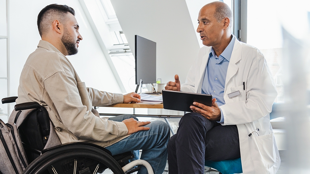 Ein Patient im Rollstuhl sitzt vor einem Arzt, der ihm mit einem iPad in der Hand etwas erklärt