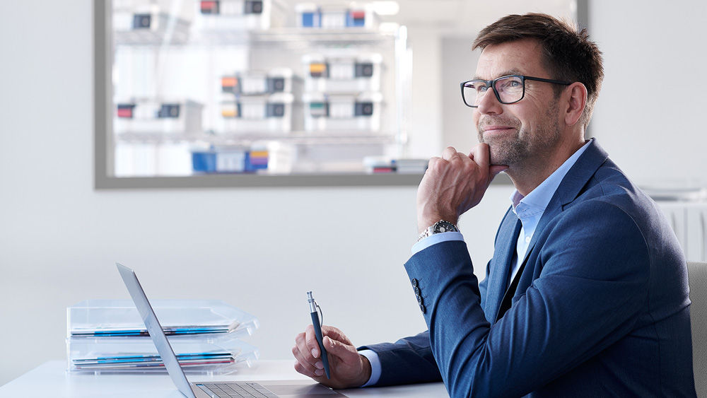 Ein Mann mit Brille sitzt in einem modernen Büro und verwendet einen Laptop mit einem sehr durchdachten Gesichtsausdruck