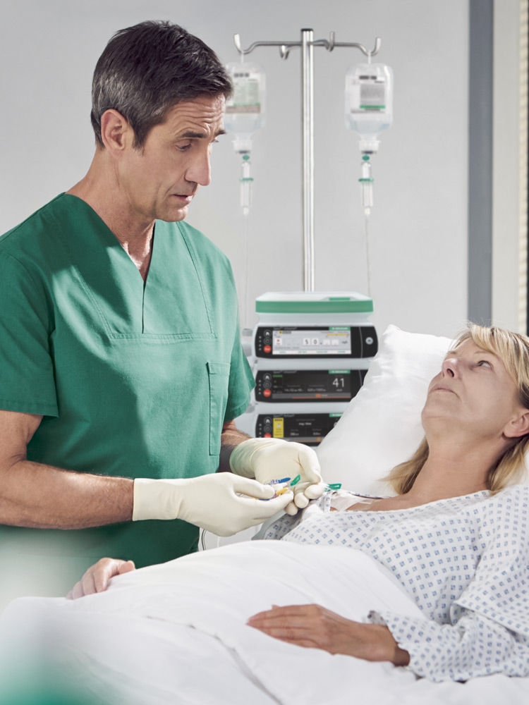 Eine blonde Frau liegt in einem Krankenhausbett. Sie ist an eine Infusion angeschlossen, im Hintergrund ist eine Spaceplus Pumpe zu sehen. Ein dunkelhaariger Arzt in einem grünen OP-Mantel steht neben dem Bett. Er reguliert die Verabreichung der Schmerzmittel per Infusion und spricht mit der Frau.