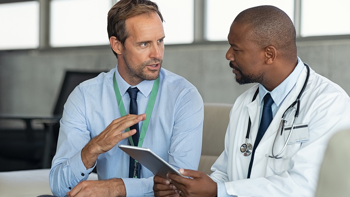 Pharmazeutischer Vertreter sitzt auf der Couch und spricht mit einem Arzt. Reifer afrikanischer Arzt bespricht die Ergebnisse der Analyse mit einem Spezialisten, während er die Diagnose auf einem digitalen Tablet berät. Arzt im Gespräch mit einem medizinischen Berater im Krankenhauszimmer, der den Fallbericht eines Patienten bespricht.