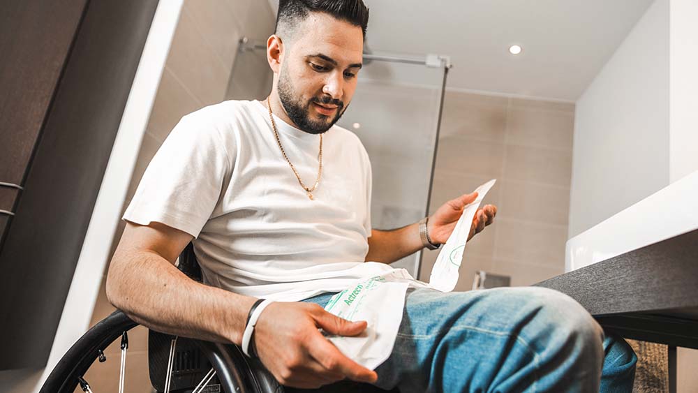 Ein Mann im Rollstuhl, ein Badezimmer im Hintergrund, hält einen Katheter in der Hand