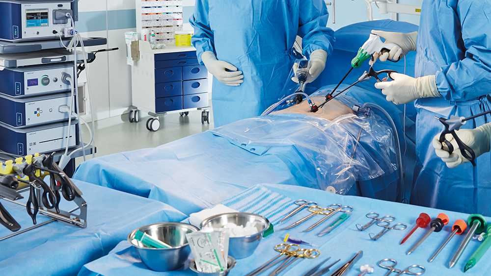 Ein vergrößertes Bild einer minimalinvasiven Operationssituation mit dem AESCULAP Caiman auf dem Bauch, mit übersichtlich angeordneten chirurgischen Instrumenten im Vordergrund; die Chirurgen beobachten die minimalinvasive Situation des Patienten auf einem AESCULAP Einstein-Monitor