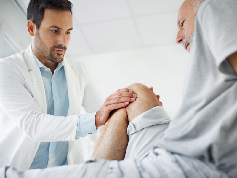 Der Patient sitzt auf einem Untersuchungstisch und die medizinische Fachkraft untersucht sanft das Knie, möglicherweise auf Schmerzen oder Mobilitätsprobleme