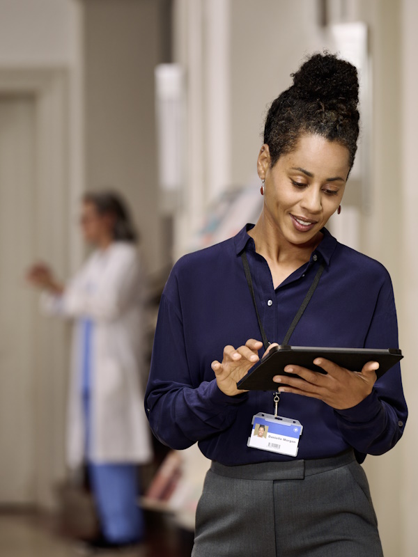 Eine Frau im Vordergrund, gekleidet in einem dunkelblauen Hemd und mit einem Ausweisschild an einem Lanyard, steht in einem Flur, hält und schaut auf ein Tablet. Im Hintergrund ist eine andere Person sichtbar, die medizinische Kleidung trägt.