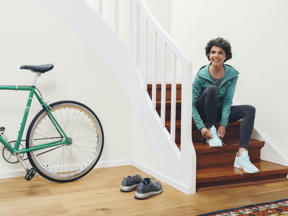 Rechts sitzt eine Frau auf der Treppe und schnürt ihre Schuhe, links ein Fahrrad und ein weiteres Paar Schuhe